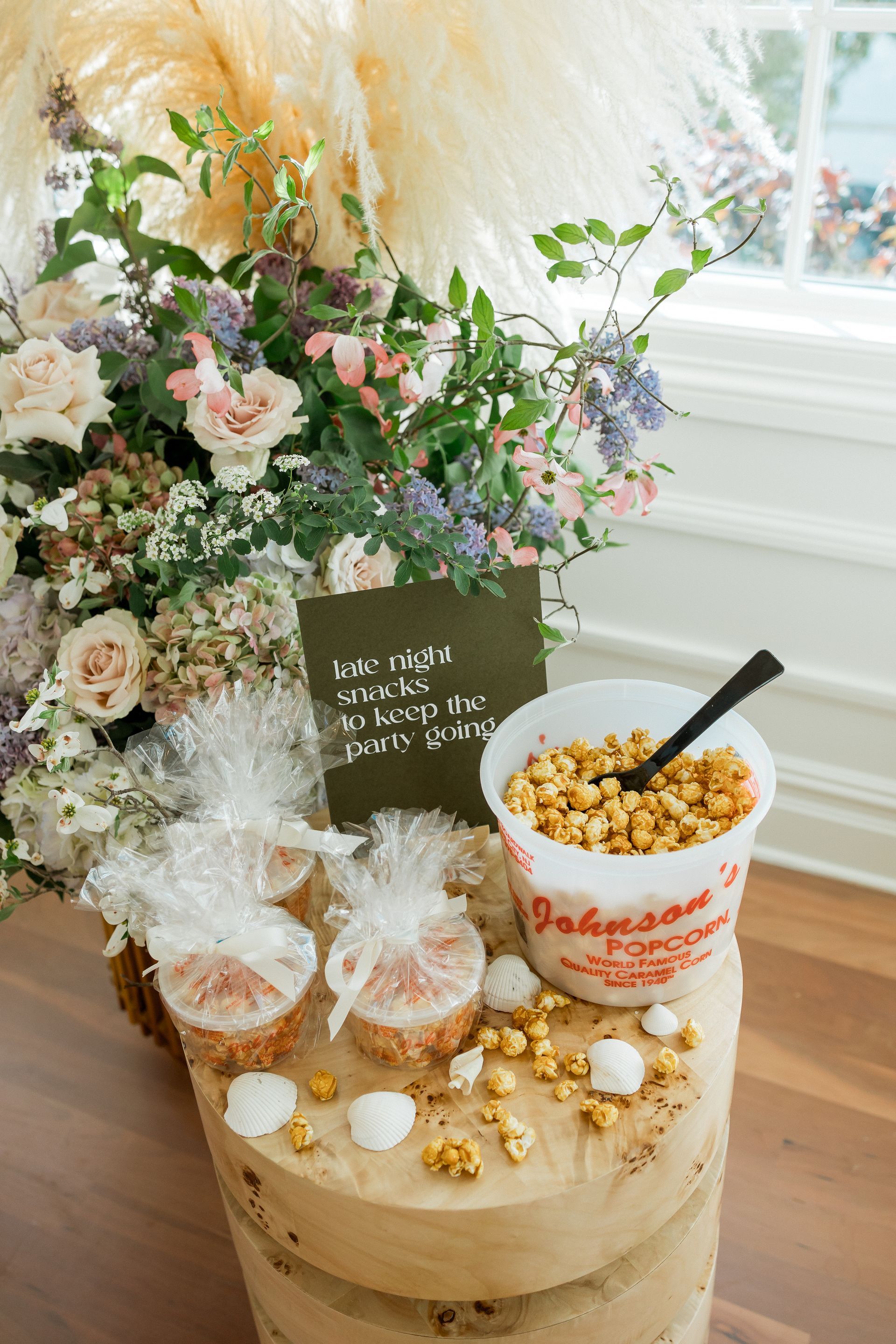Popcorn display with flowers. Bucket of caramel corn, wrapped treats, black spoon, and sign on a wood stump.