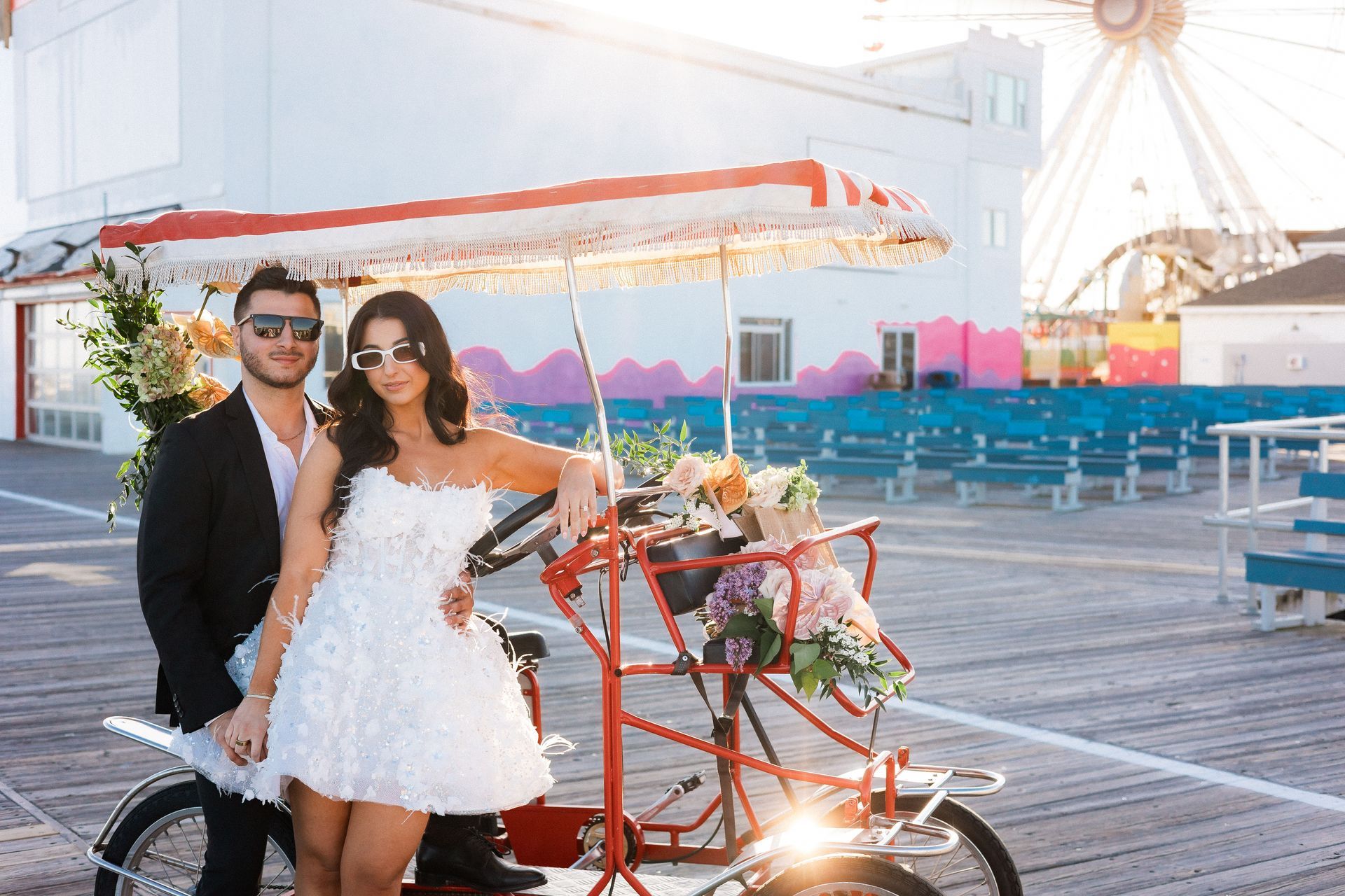 Couple posing by a red tandem bike decorated with flowers on a boardwalk, Ferris wheel in background.