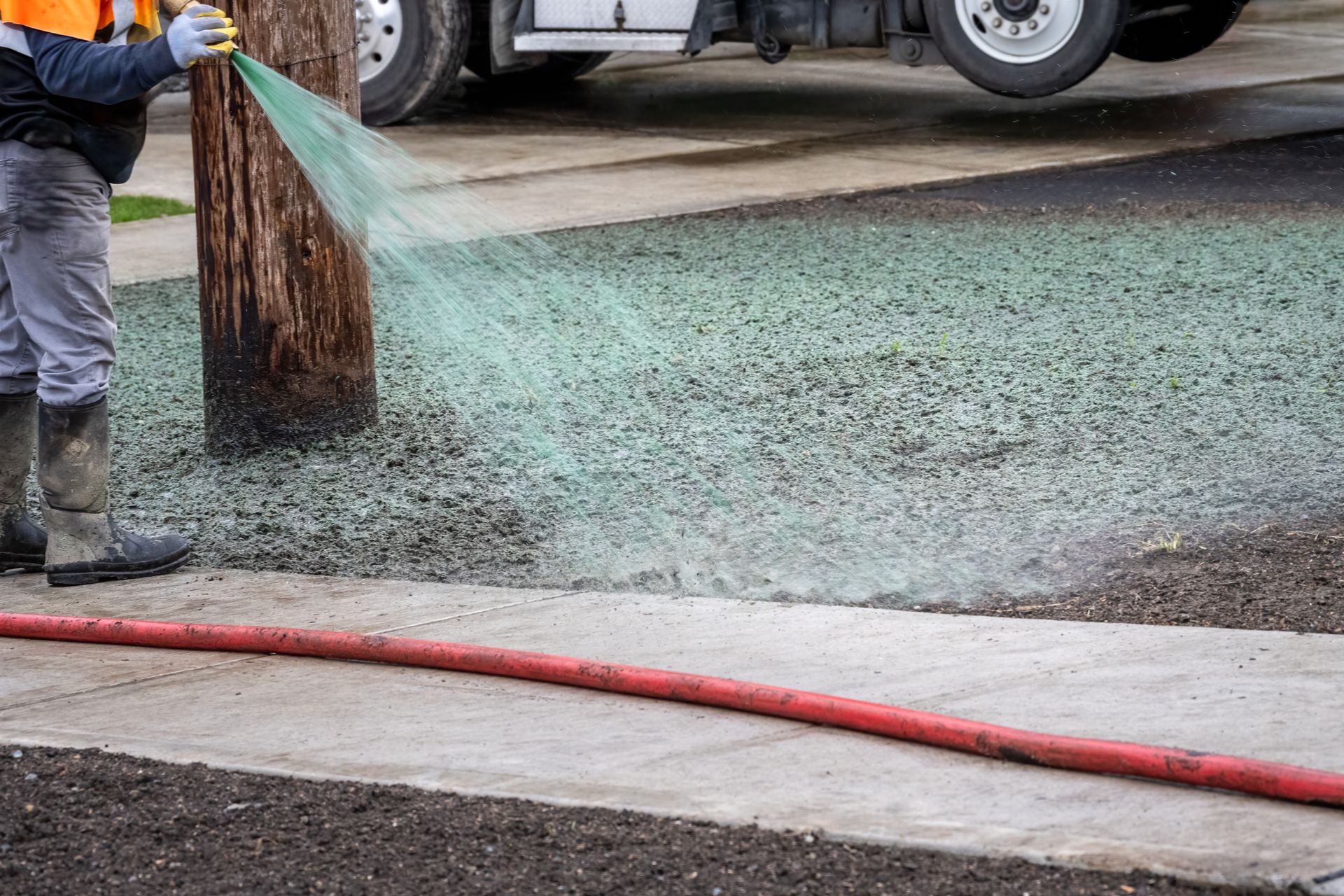 Worker spraying water over gravel at a construction site near a curb and parked truck