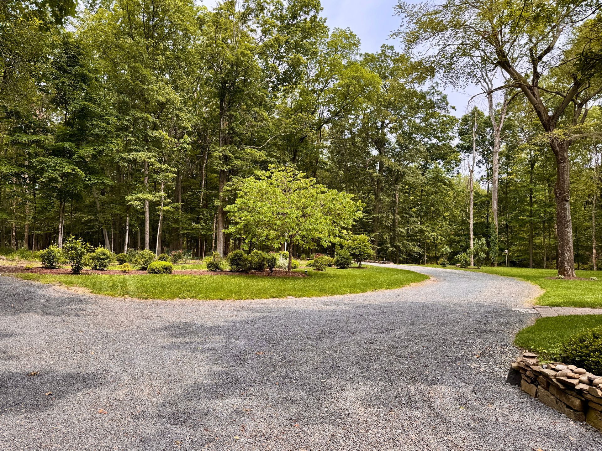 Curving gravel path through a green wooded park, with trees and stone edging on the right.