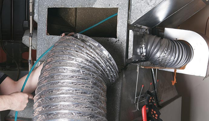 A hand vacuuming dust from a floor vent beside a blue trash bag on a hardwood floor.