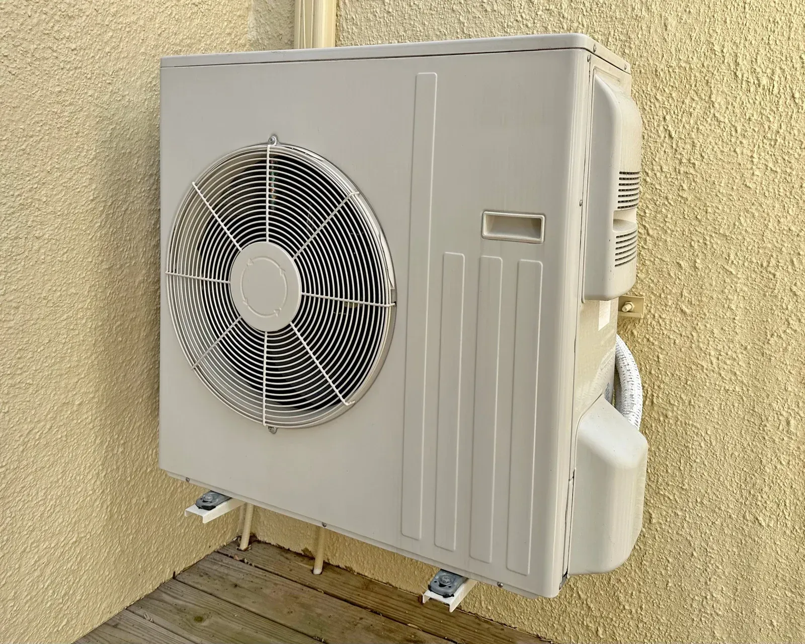 Outdoor wall-mounted air conditioner unit with a large fan on a beige stucco wall.