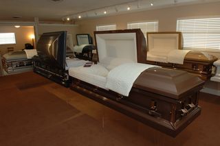 Inside a showroom, various open and closed caskets in different colors are displayed.