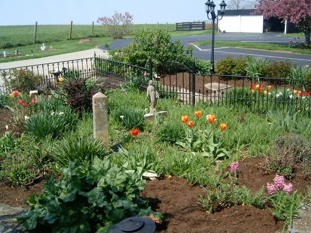 A flower garden with a cemetery in the background. Spring flowers bloom, a statue stands in the center.