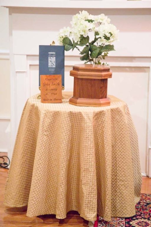 A wooden urn, flowers, a book, and a card on a table covered with a gold tablecloth.