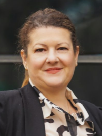 Woman with brown hair in front of a building, wearing a blazer and patterned shirt, smiling.