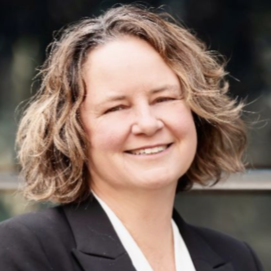 Woman with short, wavy hair smiles, wearing a black blazer and white top, outside.