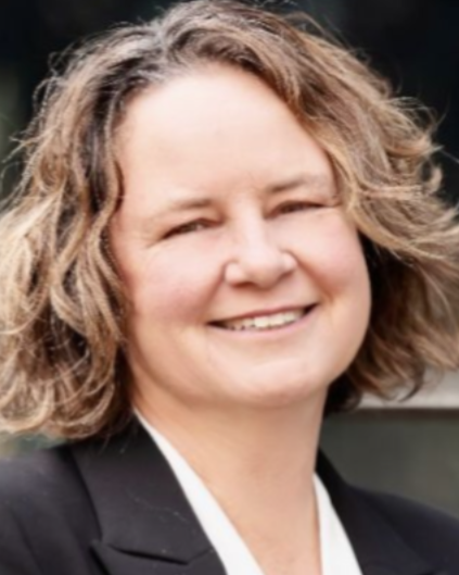 Woman smiling, wearing a black blazer and white shirt, with wavy, light brown hair.