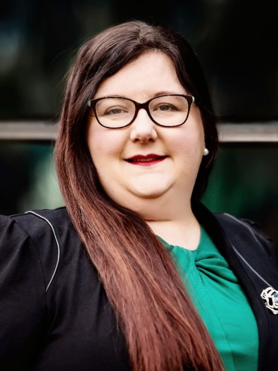 Woman with glasses, brown hair, and a green top, smiling outside.