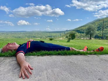 Man in blue cycling suit lies on grass, exhausted. Mountains and trees in the background, blue sky.