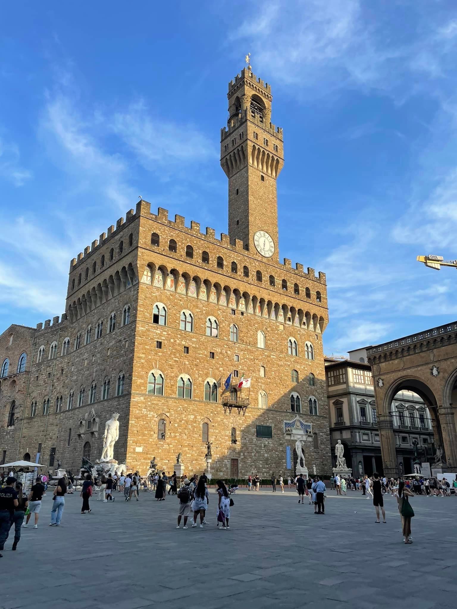 Palazzo Vecchio in Florence, Italy, with a tall clock tower, people in the piazza, and blue sky.