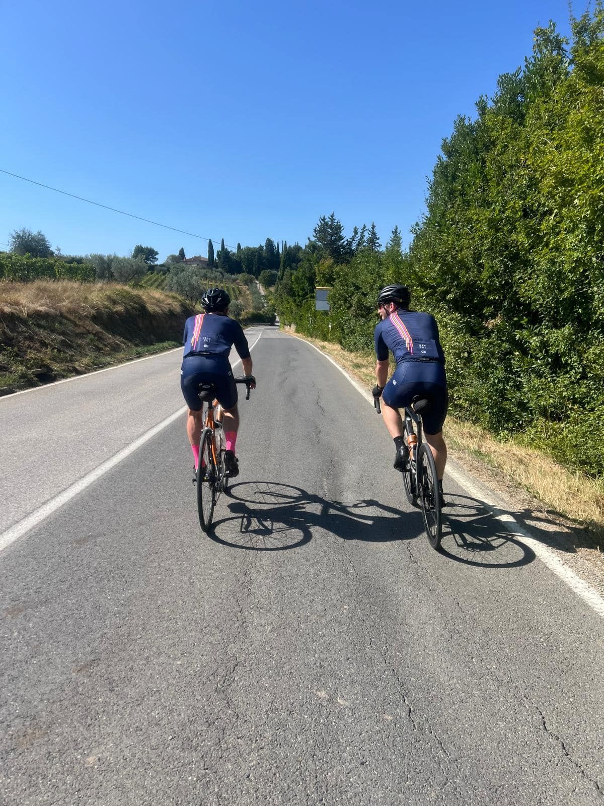 Two cyclists ride bikes uphill on a paved road under a blue sky, shadowed by trees.