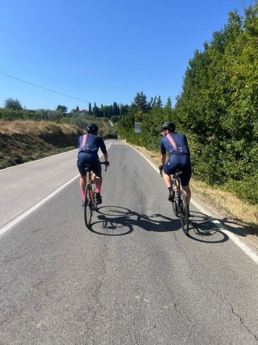 Two cyclists ride bikes uphill on a paved road under a blue sky, shadowed by trees.