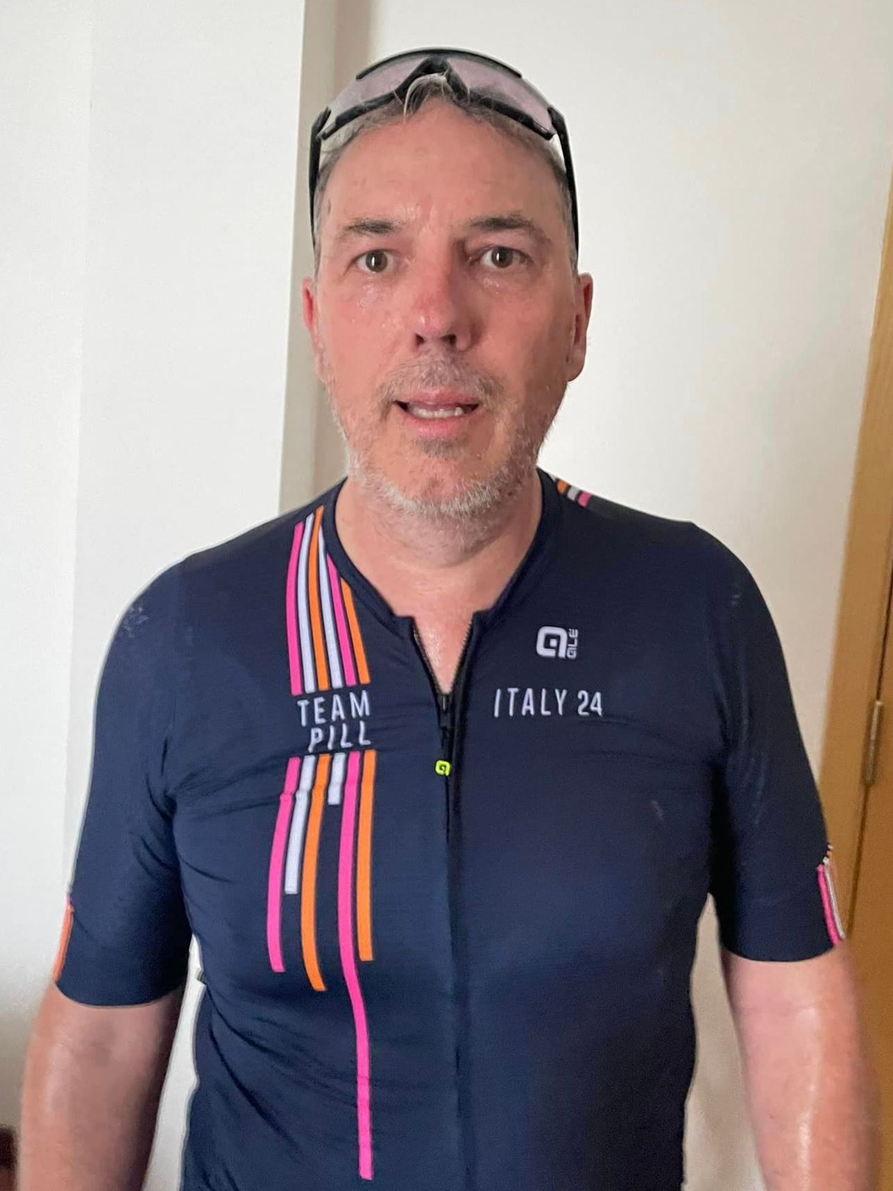 Man in cycling gear, sweating, wearing sunglasses, standing indoors. Dark blue jersey with 
