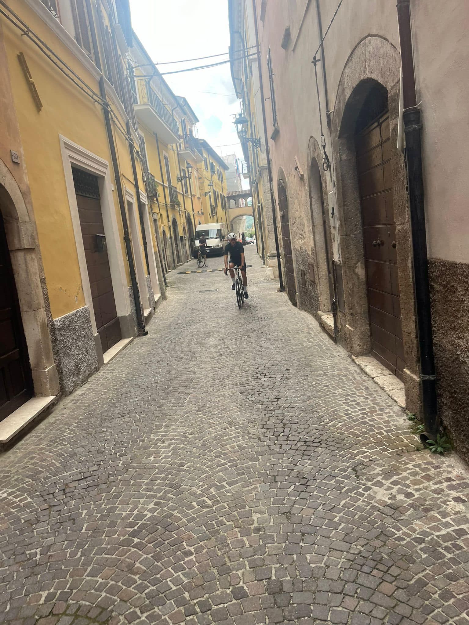 Cyclist rides down a cobblestone street in an old European town, flanked by tall buildings.