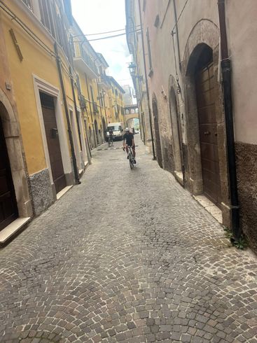 Cyclist rides down a cobblestone street in an old European town, flanked by tall buildings.
