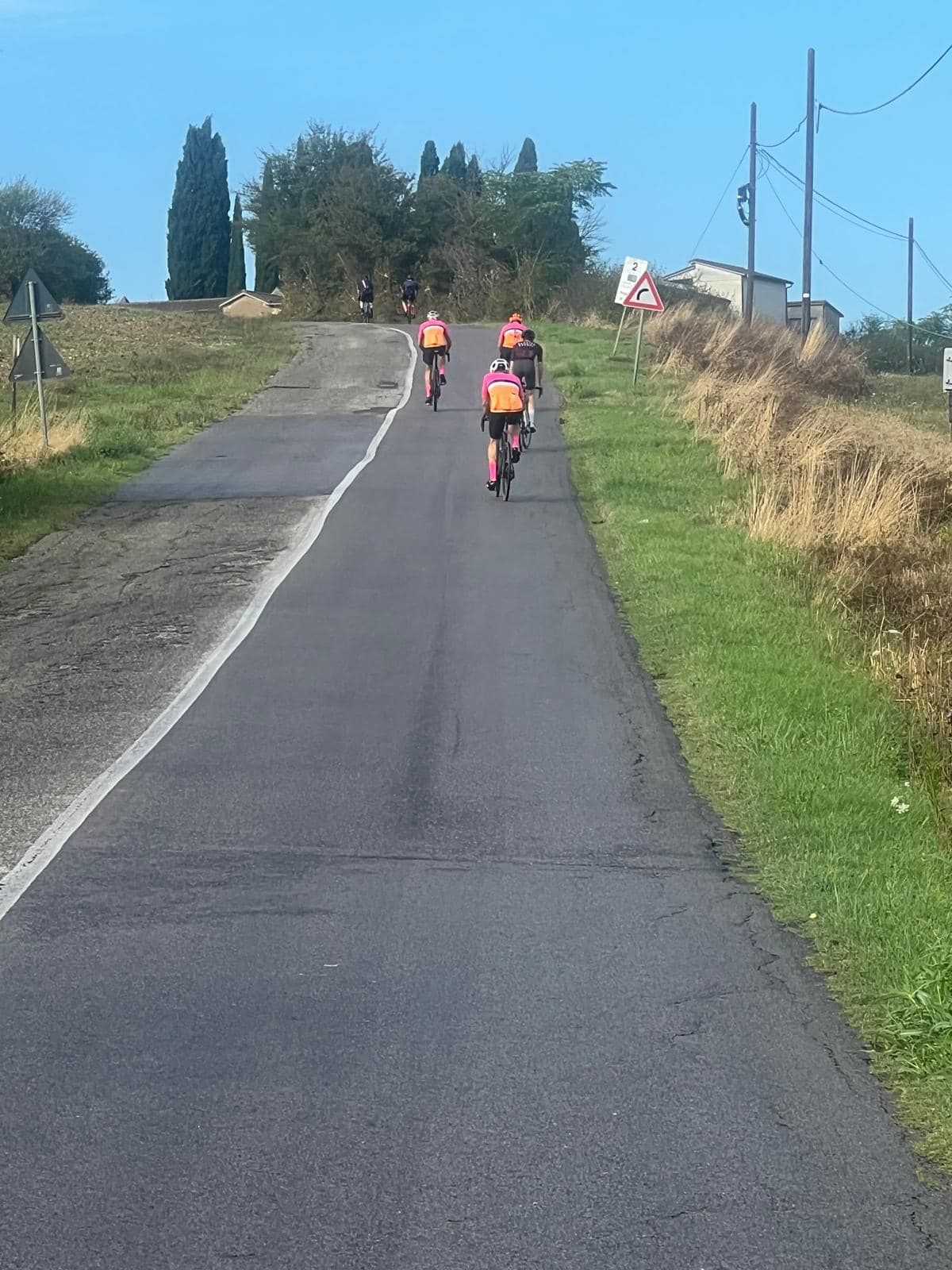 Three cyclists ride uphill on a paved road, flanked by grass and fields under a blue sky.