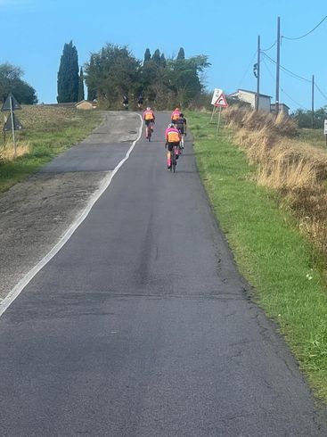 Three cyclists ride uphill on a paved road, flanked by grass and fields under a blue sky.