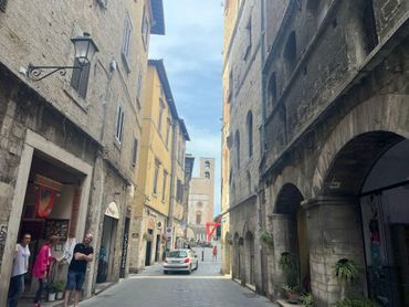 Narrow cobblestone street lined with old stone buildings, some with arched walkways. A few people and a car are present.