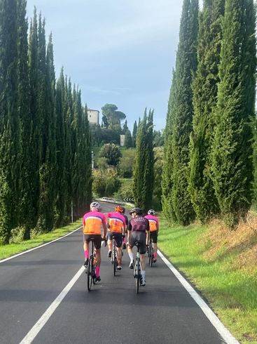 Cyclists ride down a tree-lined road with a building in the distance under a partly cloudy sky.