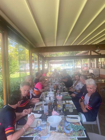 People seated at a long table, likely outdoors, eating together. Some wearing cycling gear.