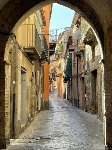 Narrow cobblestone street in an arched passage, lined with aged buildings in warm tones.