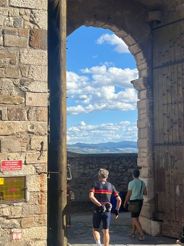 Two people walk through a stone archway, overlooking a distant city and cloudy blue sky.