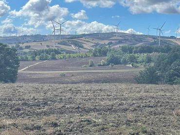 Rolling hillside with wind turbines under a cloudy sky; in the foreground, a plowed field.