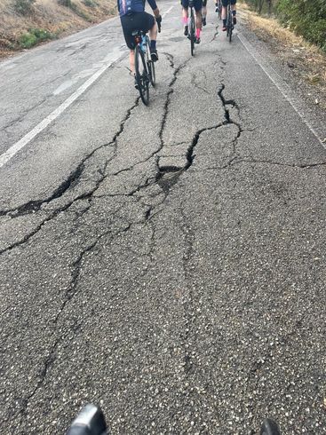 Cyclists riding on a cracked, crumbling road. Asphalt is severely damaged with large fissures.