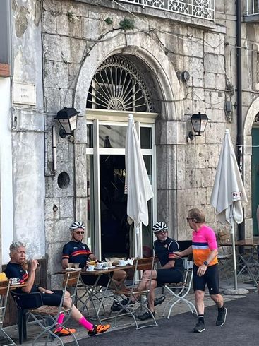Cyclists in colorful jerseys at a cafe, sitting and standing outside a stone building with arched doorway.