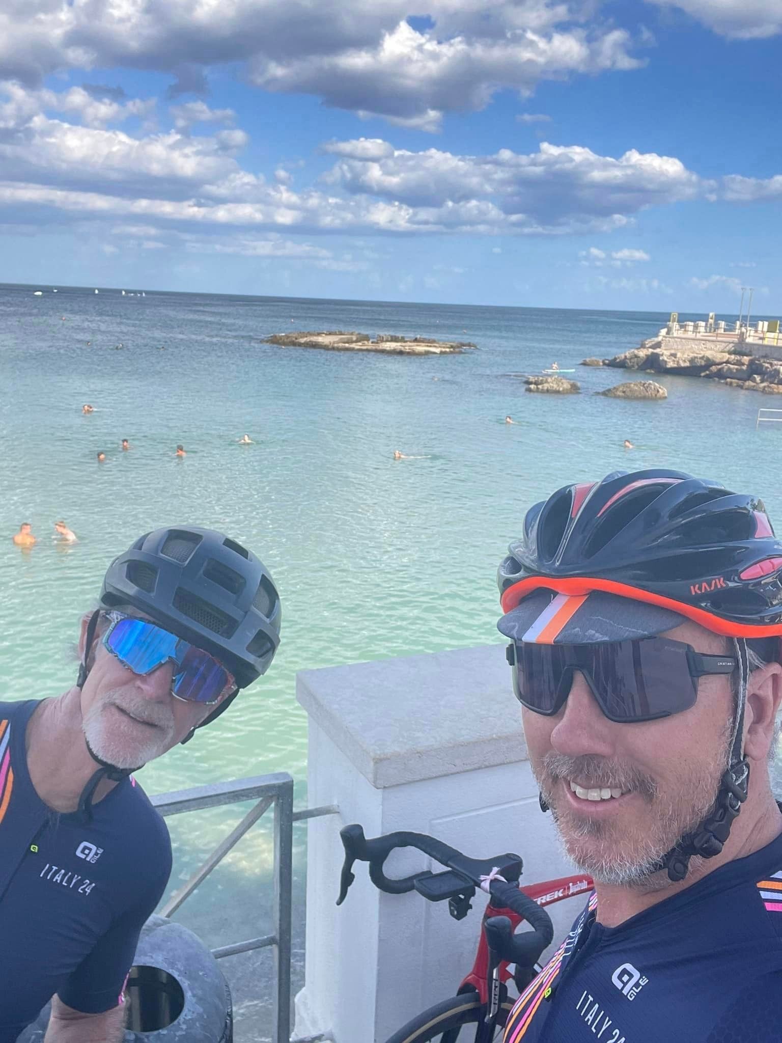 Two cyclists pose on a pier overlooking the sea; one takes a selfie. Sunny day.