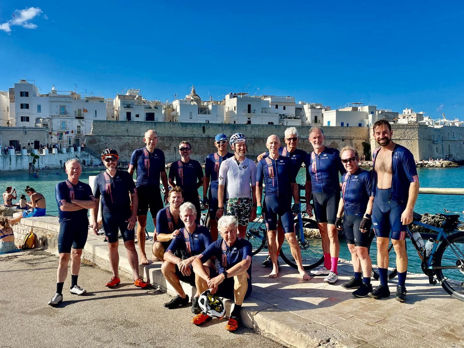 Group of cyclists in dark blue attire pose on a harbor dock against a white seaside town under a sunny sky.