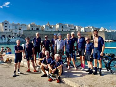 Group of cyclists in dark blue attire pose on a harbor dock against a white seaside town under a sunny sky.