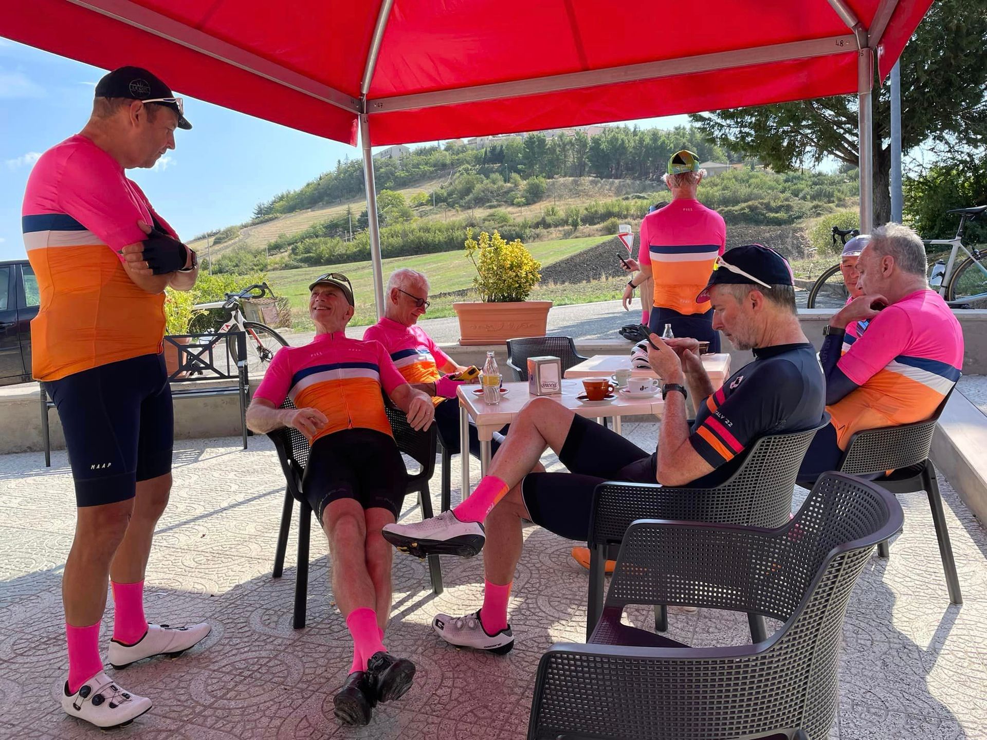 Cyclists in pink and orange jerseys relax under a red canopy at an outdoor cafe.
