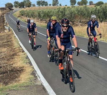 Cyclists in blue and red jerseys ride on a sunny road, passing a field.