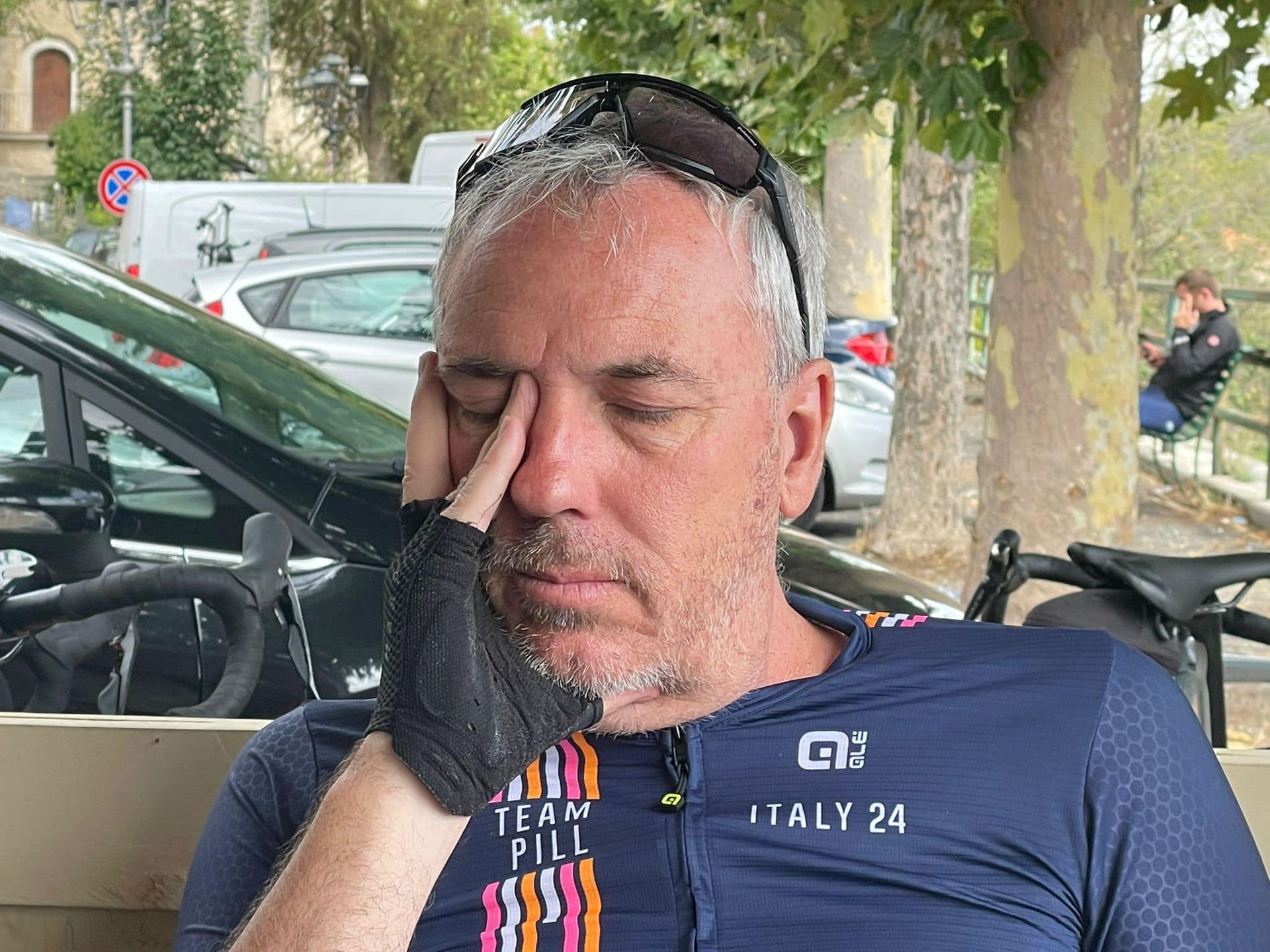Tired cyclist with gray hair, sunglasses, and glove, rubs eye, resting outside near bike.