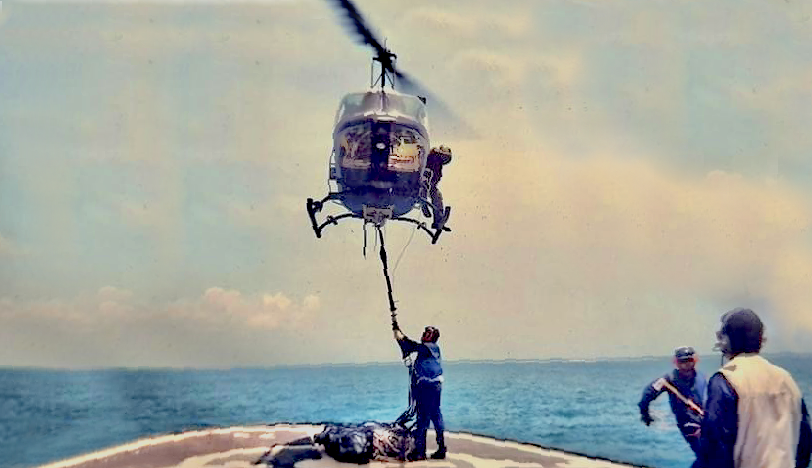 Helicopter hoisting a stretcher with a person from a ship in the ocean. Crew members watch.