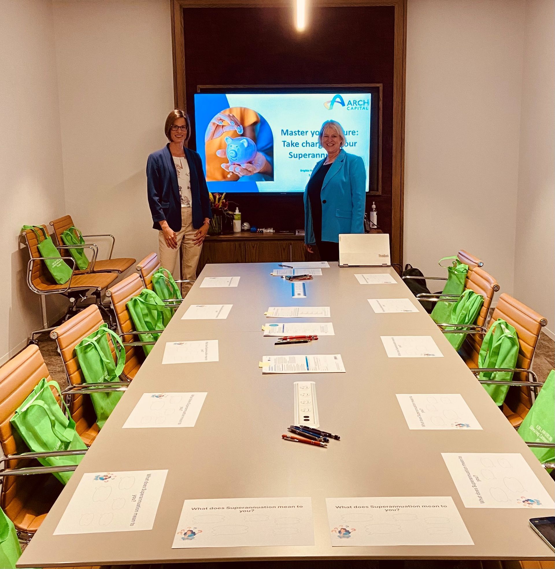 Two women stand in a conference room with a screen displaying a presentation. Papers and pens are on a long table.