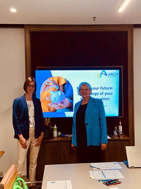 Two women stand in front of a screen; one in a blue blazer, the other in a white shirt and blazer. The screen displays a company logo and tagline.