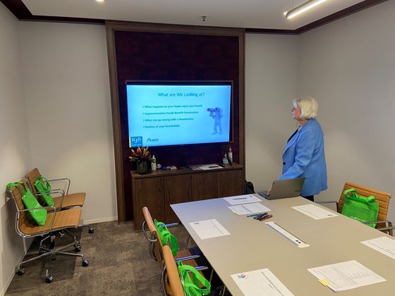 A woman in blue jacket presenting in a conference room with a screen displaying text and a graphic.