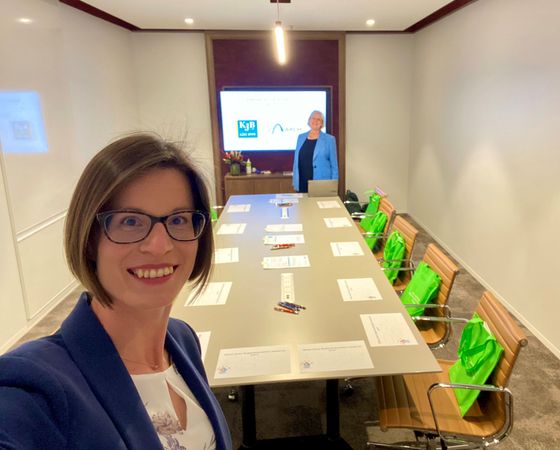 Woman in glasses takes a selfie in a conference room, with another woman standing behind the table.