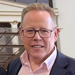 Man with glasses smiles, wearing a blazer and pink shirt, indoors.