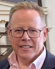 Man wearing glasses, pink shirt, and suit jacket smiles. Indoor setting, possibly office.