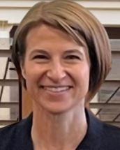 Woman with short, light brown hair smiles, dark blazer, indoors.