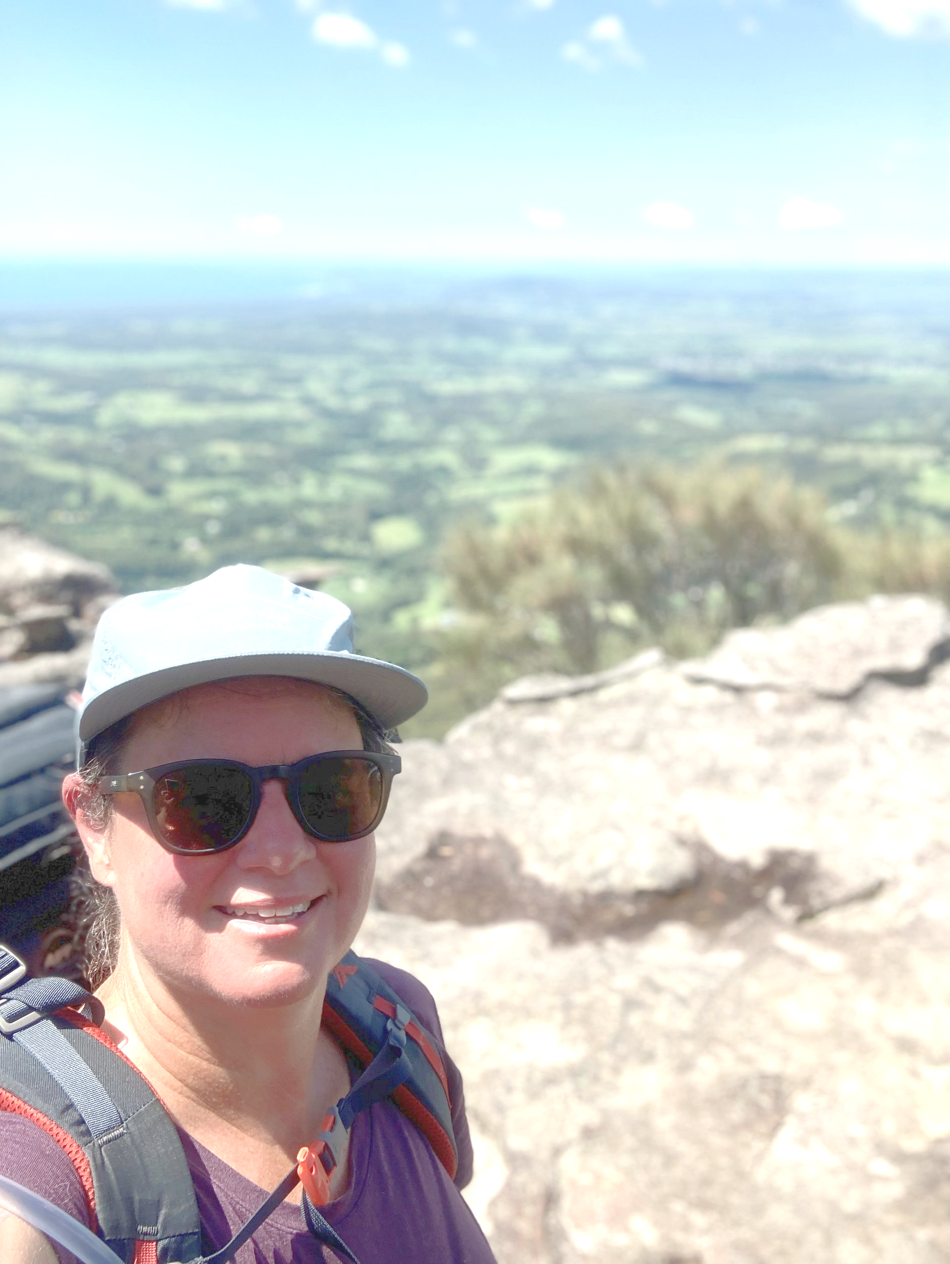 Woman wearing sunglasses and a hat smiles on a mountaintop, overlooking a green valley.