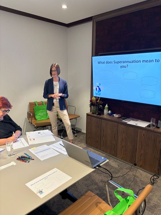 Woman giving a presentation in a meeting room, with a projector screen and a participant taking notes.