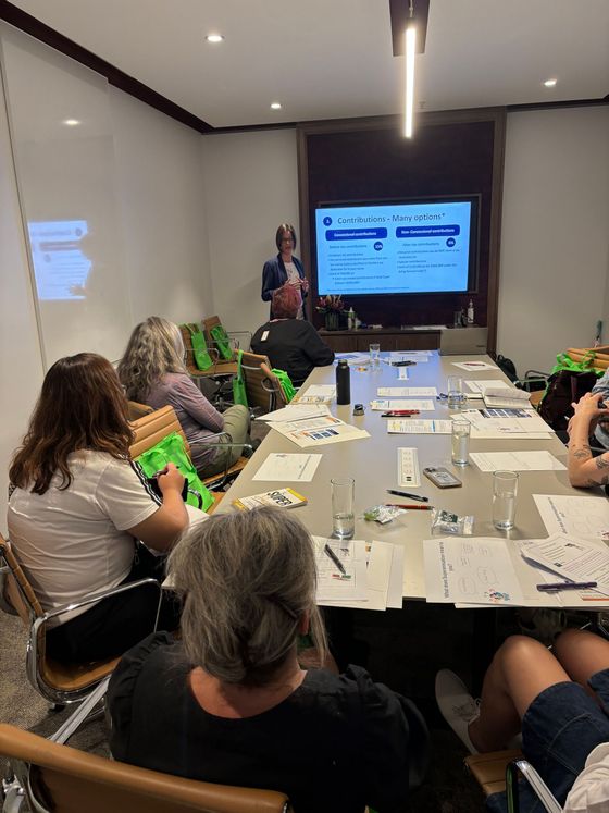 A group of people at a meeting, with a person presenting in front of a screen.
