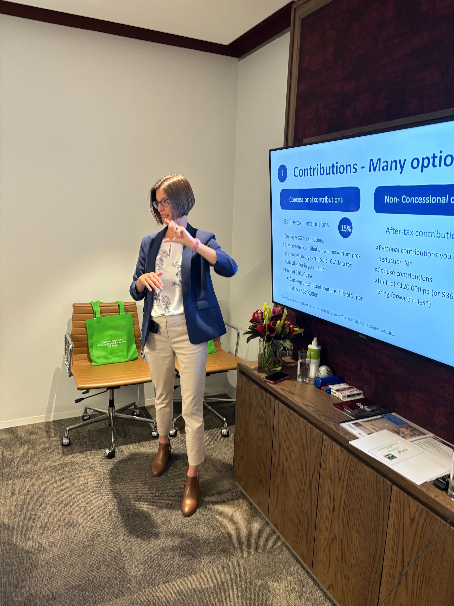 Woman presenting in a room, pointing to screen with bullet points. Beige pants, blue blazer.