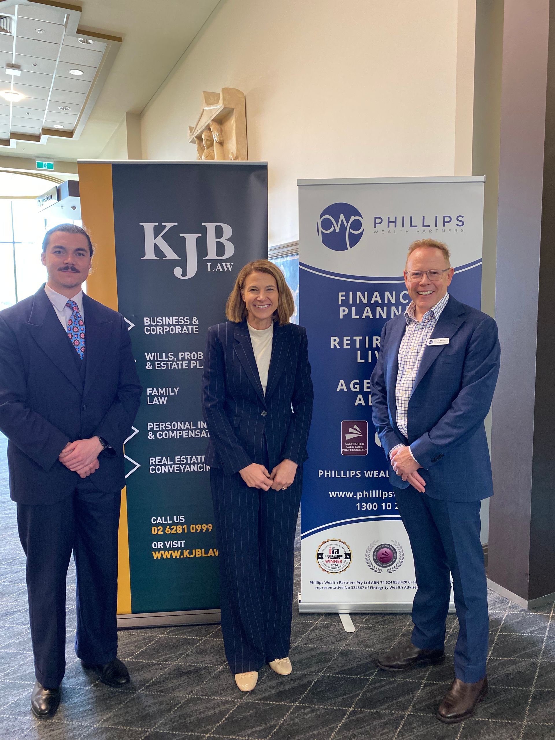 Three people standing by banners at an event, suited in navy and business casual.