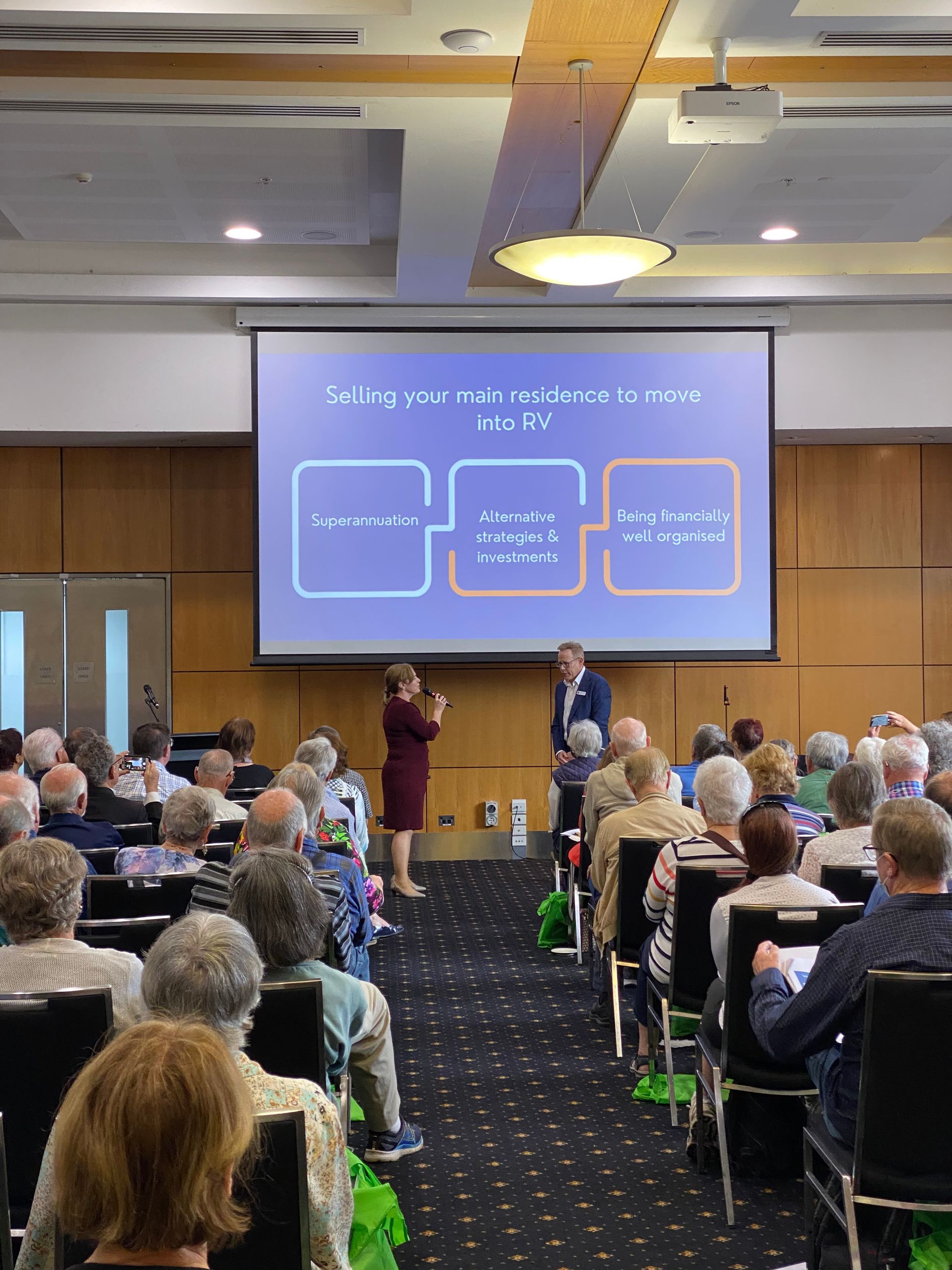 A presentation in a large conference room. Two speakers on stage address an audience of seated attendees.